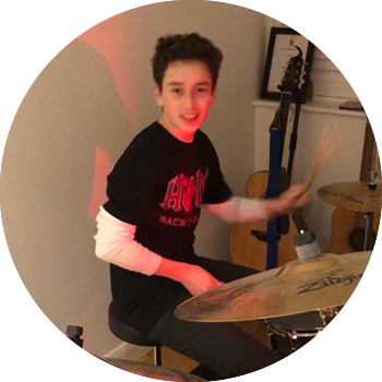 A young boy sitting at a drum kit with drumsticks in hand. The boy has dark brunette hair and is wearing a black ACDC tshirt with white sleeves. There is also two guitar in the background, of a small white room in a home.