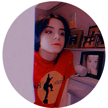 A photograph taken by the subject in the mirror freehand, they're wearing an orange graphic tshirt, they have short dark hair, there is white drawers and artwork on shelves behind them. 