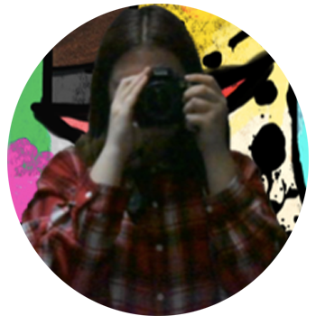 A young lady with brunette hair, with a camera covering her face, she is taking the image in the mirror, she is wearing a red and white plaid shirt. There is a colorful background. 