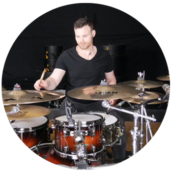 A man wearing a black t-shirt is playing on a drumkit, with a black background.