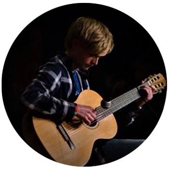 A young man sitting in the dark with a spotlight on him, an oak acoustic guitar on his lap.